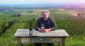 Man with a gray beard sits at a weathered wooden table on a vineyard hill, holding a glass of white wine with a bottle beside him on the table.